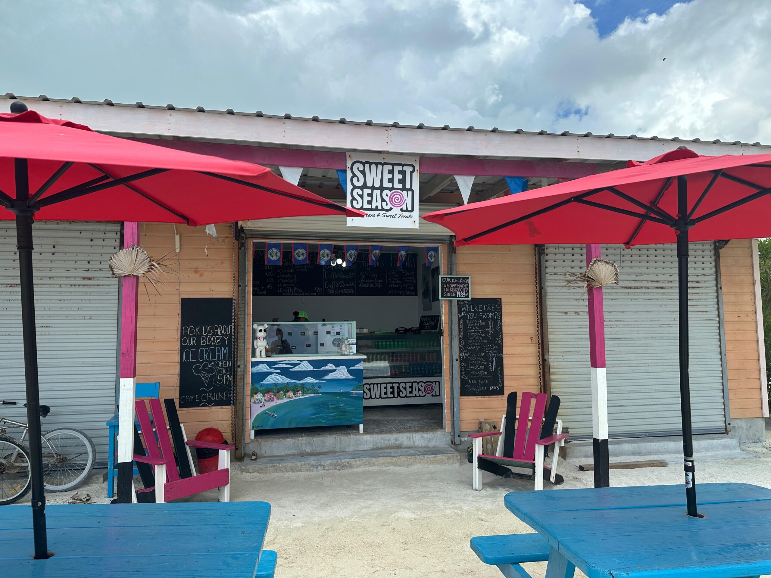 A small beachside ice cream shop called “Sweet Season,” with a colorful and laid-back vibe. The shop has wooden walls, a bright blue counter painted with a beach scene, and chalkboard menus. Outside, there are red umbrellas shading blue picnic tables and pink and blue chairs, creating a vibrant and casual atmosphere. A sign near the entrance promotes boozy ice cream, and the shop is located in Caye Caulker, Belize. The sky is partly cloudy, and bicycles are parked to the side.